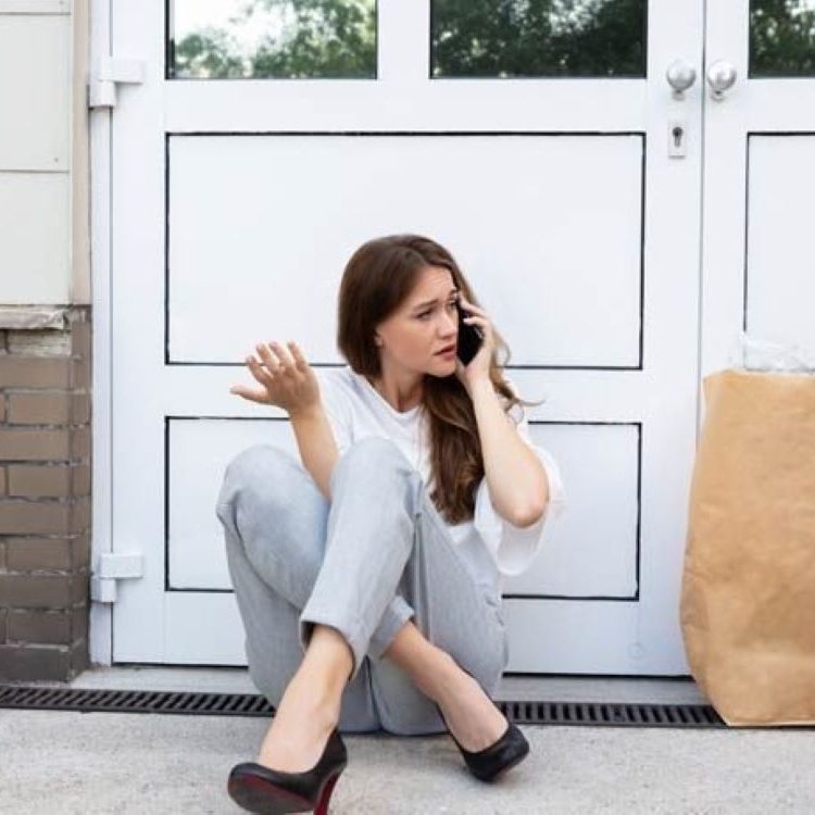 Eine Frau mit Handy sitzt vor einer Tür und schaut nachdenklich.